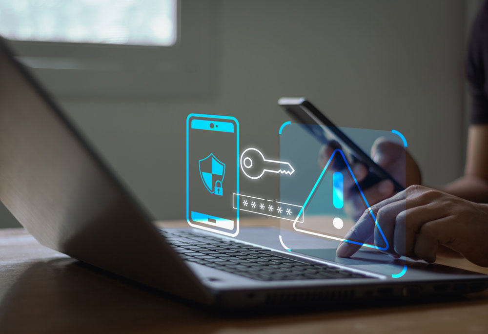 Person's hands holding a smart phone and working on a laptop with phone, password, and alert icons floating above keyboard